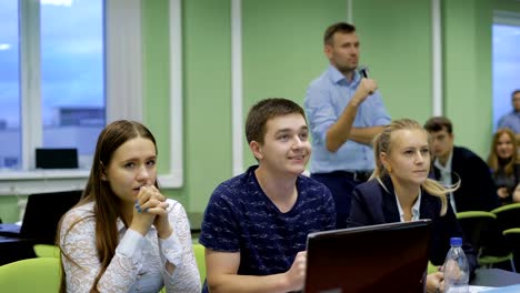 Schüler-in-der-Klasse-während-des-Unterrichts.-Lehrer-mit-Mikrofon-steht-im-Hintergrund.-Studenten-suchen-sorgfältig-vor.-Laptop-auf-dem-Tisch