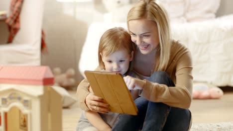 Beautiful-Mother-and-Her-Little-Daughter-Have-Good-Time-Reading-Children's-Books-on-a-Tablet-Computer.-Children's-Room-is-full-of-Toys.