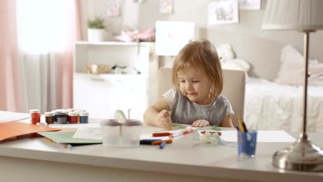 Linda-niña-se-sienta-en-su-mesa-y-dibuja-con-crayones.-Su-habitación-es-rosa,-dibujos-bonitos-colgando-de-las-paredes,-muchos-juguetes-por-ahí.