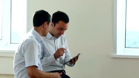 Two-business-executives-sitting-in-a-bright-office-space,-looking-for-information-together-by-sharing-the-screen-of-a-digital-tablet