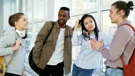 Dolly-shot-view-of-four-male-and-female-student-of-differnt-ethnicity-standing-near-window-in-wide-white-hallway-discussing-study-amiling-and-laughing