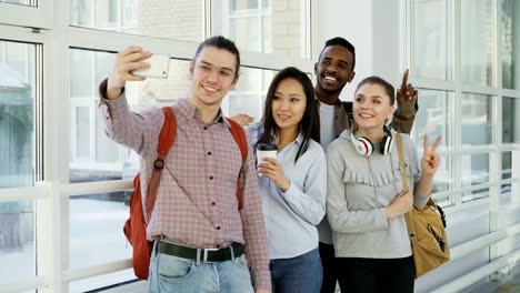 Group-of-four-multi-ethnic-positive-male-and-female-students-are-standing-in-wide-corridor.-Hipster-gut-is-holding-smartphone-making-selfie-of-them-all