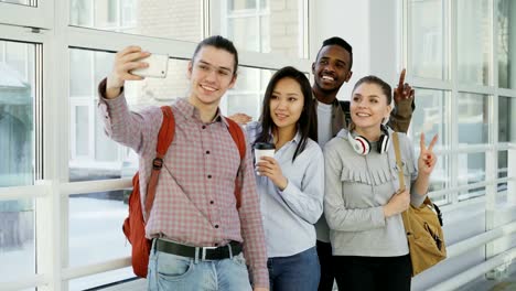 Grupo-de-cuatro-estudiantes-hombres-y-mujeres-positivos-multiétnicos-está-de-pie-en-el-amplio-corredor.-Tripa-de-hipster-tiene-smartphone-haciendo-selfie-de-todos