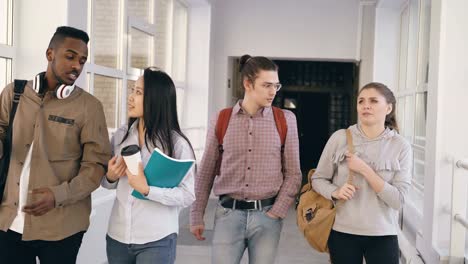 Vier-Kollegen-unterschiedlicher-ethnischer-Herkunft-sind-Fuß-in-lange-geräumigen-weiten-gläsernen-Korridor-in-Universität-Lächeln-und-lachen-und-reden-miteinander