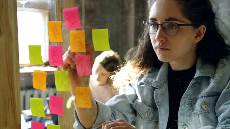 Close-up-of-young-creative-entrepreneur-numbering-coloured-stickers-on-glassboard-while-working-together-with-female-colleague-in-modern-office