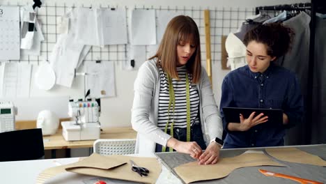 Weibliche-Kleidung-Designer-ist-Umrisse-der-neuen-Gewand-auf-Stoff-mit-Kreide-zeichnen,-während-ihr-Kollege-ihr-hilft-und-Tablet-verwenden.-Technologien-in-der-Kleidung-Herstellung-Konzept.