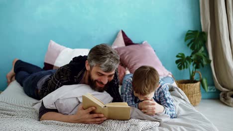 Caring-father-is-reading-funny-book-to-his-child-while-boy-is-laughing-and-talking-to-his-parent.-Full-size-bed,-bright-pillows-and-green-plants-are-visible.