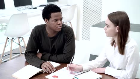 Two-economics-students---Caucasian-woman-and-African-man-discussing-document-with-graphs-and-charts.-Classmates-sitting-at-desk-and-preparing-project-together