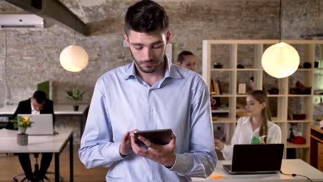 Young-businessman-is-surfing-internet-on-tablet-in-office,-watching-at-camera,-his-colleagues-are-networking-with-technologies,-working-concept,-business-concept,-communication-concept