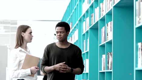 Two-college-students,-Caucasian-woman-and-African-man-standing-by-bookshelves-in-library-and-discussing-books