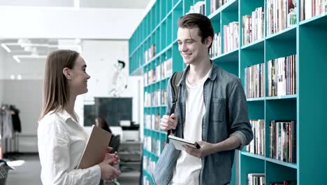 Dolly-in-shot-of-two-university-students,-boy-and-girl,-having-friendly-talk-near-bookshelves-in-library