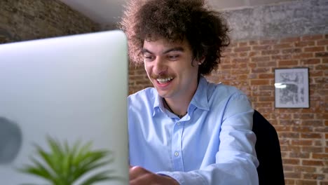 Happy-businessman-with-curly-large-hair-cheering-about-victory,-winner-sitting-at-table-with-laptop,-modern-office-background