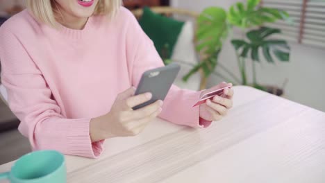 Beautiful-Asian-woman-using-smartphone-buying-online-shopping-by-credit-card-while-wear-sweater-sitting-on-desk-in-living-room-at-home.-Lifestyle-woman-at-home-concept.