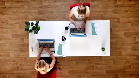 Top-down-shot,-two-hard-working-females-typying-on-their-laptops-and-sitting-at-the-table-in-the-wooden-studio