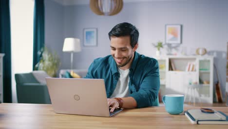 Mittlere-Aufnahme-ein-hübscher-Mann-sitzt-an-seinem-hölzernen-Schreibtisch-arbeiten-auf-einem-Laptop-in-seinem-gemütlichen-Wohnzimmer.