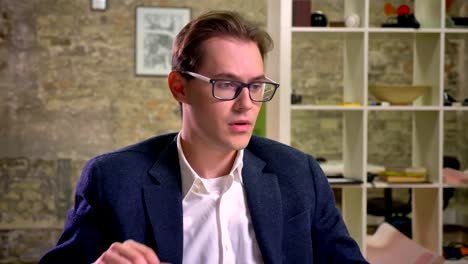 Cute-businessman-with-caucasian-appearance-is-fixing-his-glasses-and-looking-through-them-at-camera-with-happy-smile,-sitting-at-white-desk-in-office