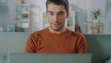 Handsome-Man-Uses-Laptop-Computer-while-Sitting-on-Sofa-at-Home.-Man-Working,-Browsing-Through-Internet-from-His-Cozy-Living-Room.