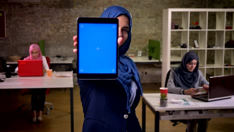 Linda-mujer-agradable-en-hijab-azul-muestra-pantalla-azul-y-tableta-de-explotación-a-la-cámara,-en-ladrillo-luz-oficina-junto-a-otras-chicas-de-Oriente