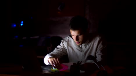 Shooting-of-dark-office,-light-from-screen-ar-shining-on-his-face-while-he-is-sitting-looking-at-computer-and-checking-papers,-holding-them-with-focused-confident-face,-indoor-illustration