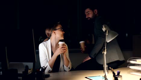 Caring-male-colleague-bearded-man-is-bringing-take-away-coffee-to-tired-young-woman-working-on-computer-late-at-night.-Girl-and-guy-are-talking-and-smiling.
