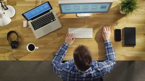 Vista-superior-de-un-joven-ingeniero-trabajando-en-su-computadora-de-escritorio-y-Laptop.-Filmada-en-una-hermosa-mañana-de-verano,-la-luz-del-sol-cae-sobre-su-mesa-de-madera.