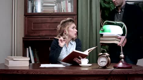 Concentrated-young-woman-reading-a-book-with-a-pencil-in-her-hands,-sitting-at-a-desk-when-her-male-colleague-brings-her-another-pile-of-books