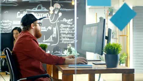 In-the-Creative-Agency-Videographer-in-a-Cap-Works-on-Personal-Computer-with-Two-Monitors,-He's-Doing-Video-and-Audio-Editing.-Stylish-Coworkers-at-Their-Workstations-and-Chalkboard-Wall-Visible.