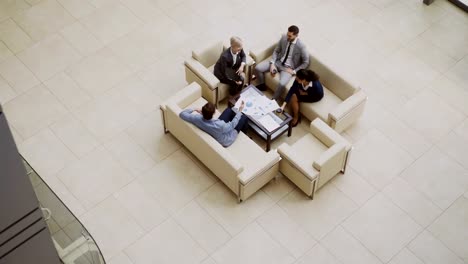 Top-view-of-group-of-business-people-colleagues-discussing-financial-charts-sitting-on-couchs-in-lobby-at-modern-business-center