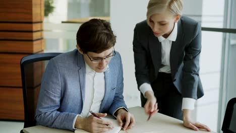 Dos-compañeros-de-trabajo-discutiendo-los-informes-en-la-oficina-moderna.-Hombre-de-negocios-sentado-en-la-mesa-hablando-a-su-pareja-femenina