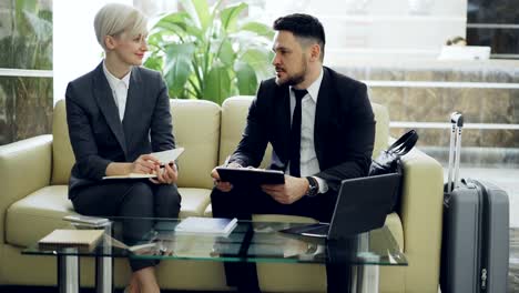 Two-female-and-male-business-colleagues-with-clipboard-and-notepad-talking-at-hotel-lobby.-Luggage-in-near-couch.-Business,-travel-and-people-concept