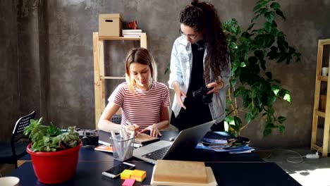 Young-creative-colleagues-are-planning-design,-drawing-layout-in-notebook-and-watching-photos-on-camera-screen.-Women-are-busy-working-on-important-project.