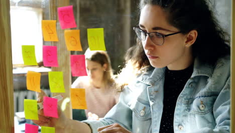 Joven-empresaria-creativa-numeración-pegatinas-de-colores-en-glassboard-proyecto-mientras-trabajaba-junto-a-colega-en-la-oficina-moderna