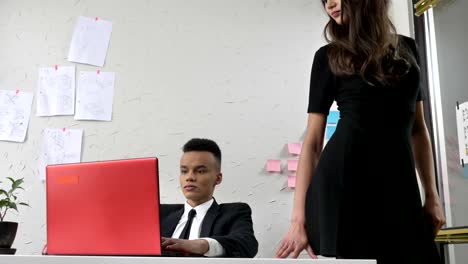 Young-woman-boss-sits-down-on-the-table-to-her-African-worker,-gently-touches-his-hand,-flirts,-harassment-concept-50-fps