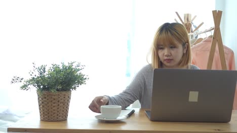 Beautiful-young-smiling-woman-working-on-laptop-while-enjoying-drinking-warm-coffee-sitting-in-a-living-room-at-home.-Enjoying-time-at-home.-Asian-business-woman-working-in-her-home-office.