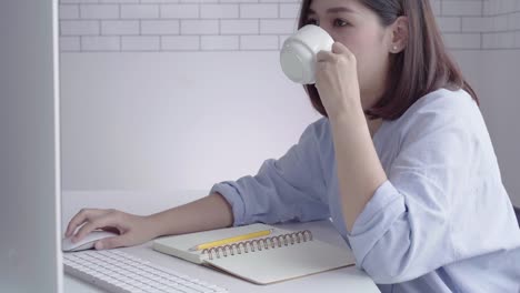 Beautiful-young-smiling-woman-working-on-laptop-while-enjoying-drinking-warm-coffee-sitting-in-a-living-room-at-home.-Enjoying-time-at-home.-Asian-business-woman-working-in-her-home-office.