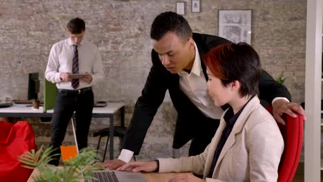 Young-asian-woman-and-black-man-talking-over-laptop-in-modern-office,-caucasian-man-standing-in-back-with-tablet