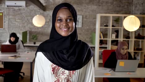 Joven-negro-a-las-mujeres-musulmanas-en-hijab-de-pie-y-mirando-en-la-cámara,-Sonriente,-tres-mujeres-trabajando-en-ordenador-portátil-en-la-oficina-moderna