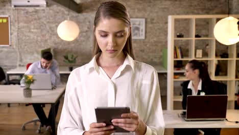 Young-concentrated-businesswoman-is-tapping-on-tablet-in-office,-watching-at-camera,-her-colleagues-are-networking-with-technologies,-working-concept,-business-concept,-communication-concept