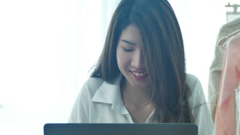 Beautiful-young-smiling-woman-working-on-laptop-while-enjoying-drinking-warm-coffee-sitting-in-a-living-room-at-home.-Enjoying-time-at-home.-Asian-business-woman-working-in-her-home-office.