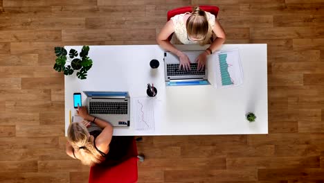 Top-down-shot,-two-productive-girls-sitting-at-the-table-in-wooden-office-and-using-their-laptops-and-blue-screen-smartphone