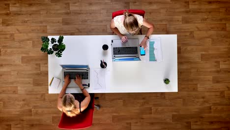 Top-down-shot,-two-working-blondes-at-wooden-office-checking-paper-graphs-with-their-screens-precisely,-blue-screen-phone-is-lying-next-to-laptop