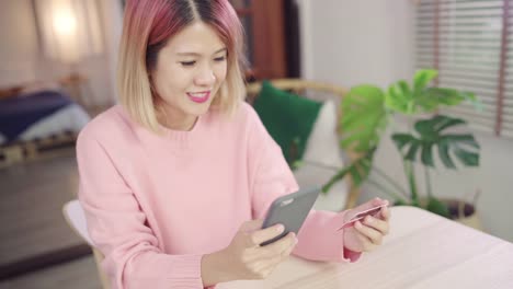 Beautiful-Asian-woman-using-smartphone-buying-online-shopping-by-credit-card-while-wear-sweater-sitting-on-desk-in-living-room-at-home.-Lifestyle-woman-at-home-concept.