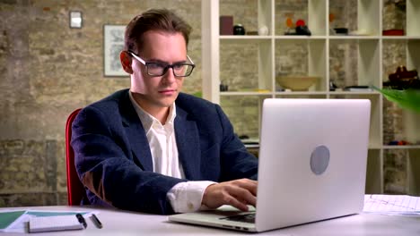 Concentrated-caucasian-male-is-sitting-and-looking-at-his-working-laptop-and-then-at-camera-relaxed-on-brick-background