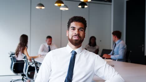 Portrait-Of-Businessman-In-Modern-Boardroom-With-Colleagues-Meeting-Around-Table-In-Background-Shot-In-Slow-Motion