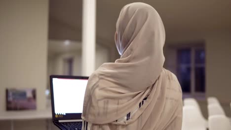 Young-arabic-female-wearing-a-hijab-going-by-the-hall-while-holding-her-black-laptop-in-hands.-Employee,-working-place,-conference-hall,-corridor.-Backside-view,-unfocused-background