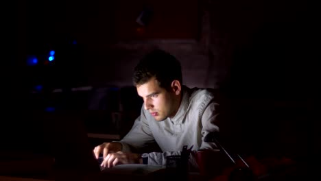 Nice-caucasian-man-is-tired-from-typing-on-laptop,-started-falling-asleep-next-to-the-screen-while-sitting-at-workplace-inside-dark-office,-black-background