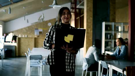 Caucasian-positive-woman-working-in-public-workplace-with-brick-wall-interior.-Beautiful-young-brunette-in-plaid-shirt-is-working-in-modern-workspace,-walking-by-room-with-laptop.-Smiling,-positive-business-woman-greets-colleagues