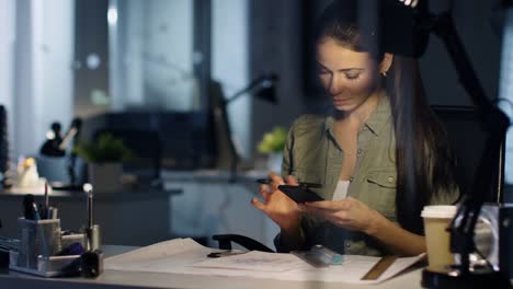 Spät-bei-Night-Female-Design-Engineer-Works-on-Drafts,-She-Uses-Smartphone-and-Makes-Corrections-on-Her-Schemes.-Office-ist-nur-von-Ihrer-Tischlampe.