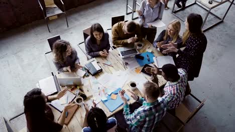 Vista-del-grupo-de-personas-de-raza-mixta-sentado-en-la-mesa,-hablando-de-superior-y-entonces-aplaudiendo-juntos.-Reunión-de-negocios