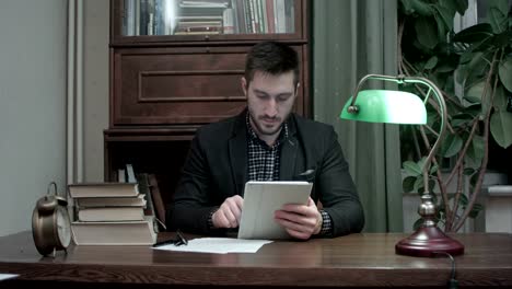 Young-man-sitting-at-desk-with-books-using-digital-tablet-in-home-room-office